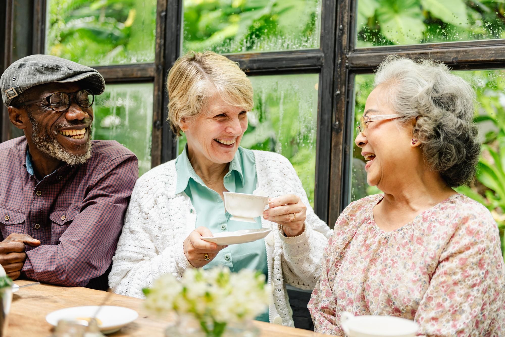 Three older adults laughing and enjoying tea together by a window with greenery outside.