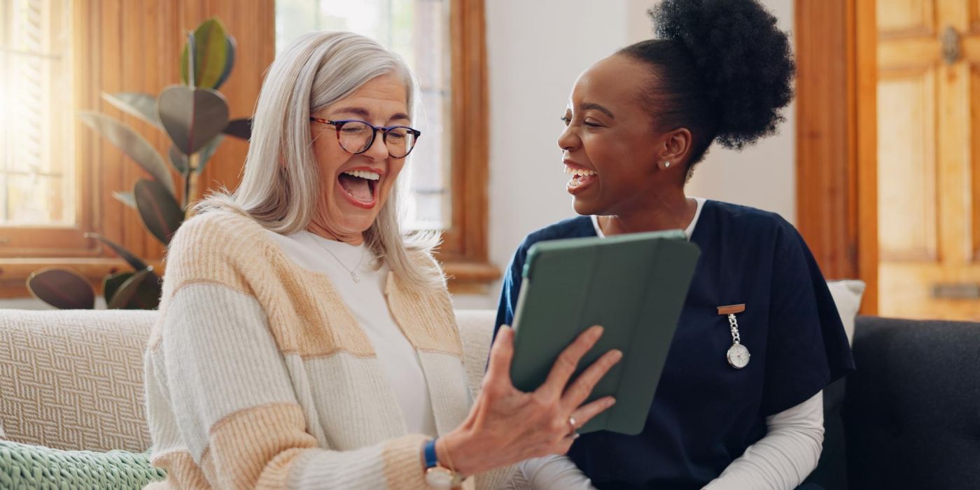 Two women smiling and laughing together while looking at a tablet on a sofa in a bright living room.