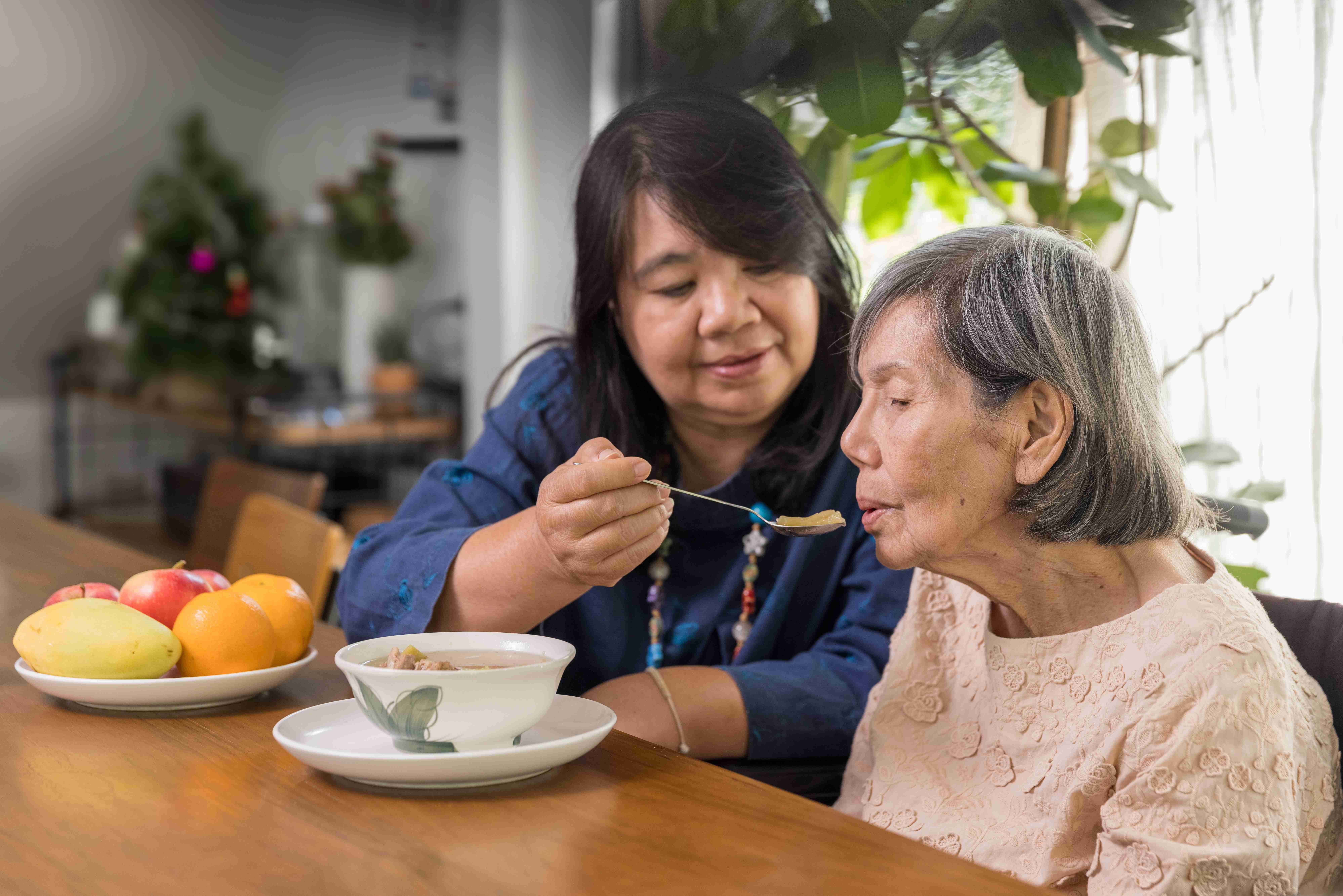 Woman feeding an elderly lady at a table with a bowl of soup and a plate of fruit.