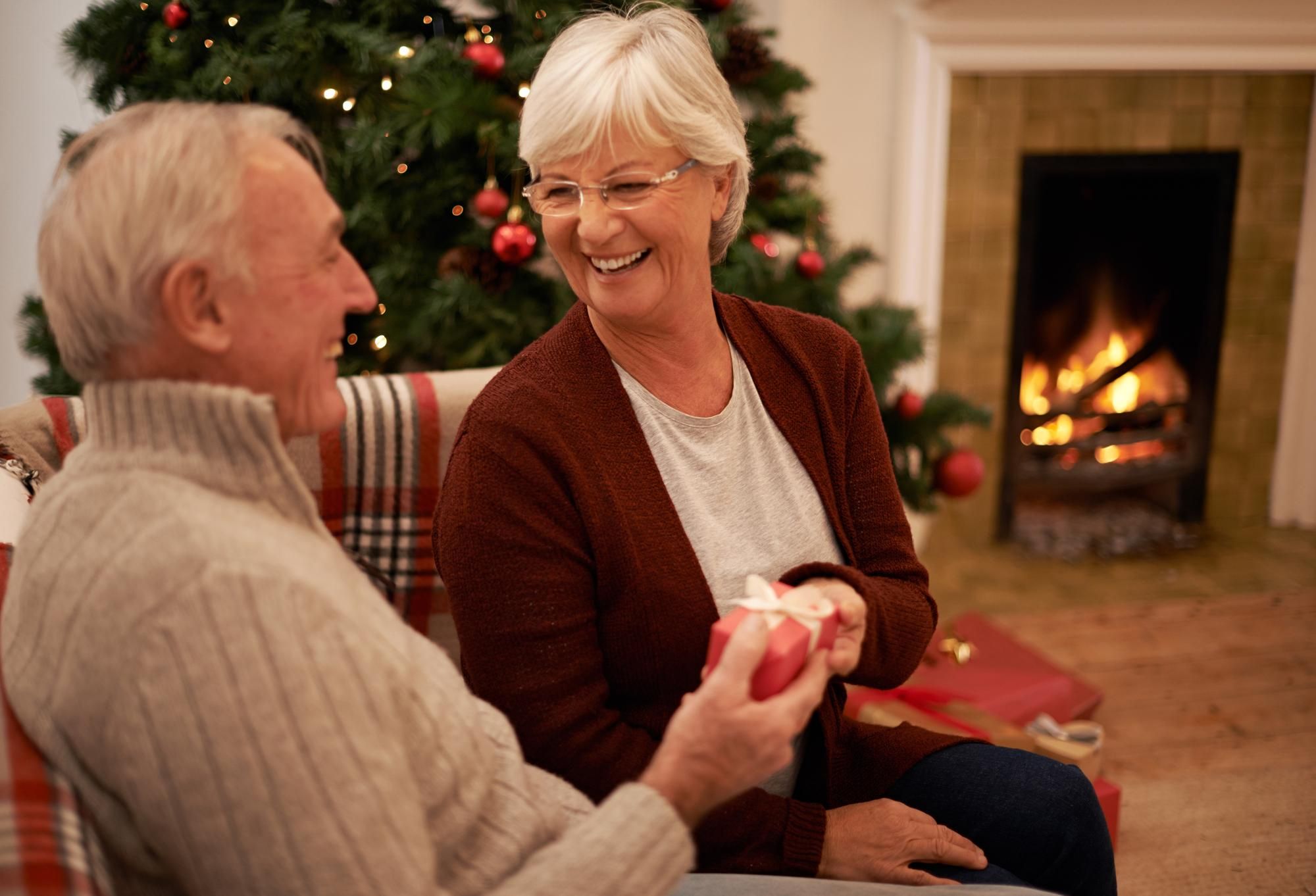 Smiling older couple exchanging a gift by a Christmas tree and fireplace.