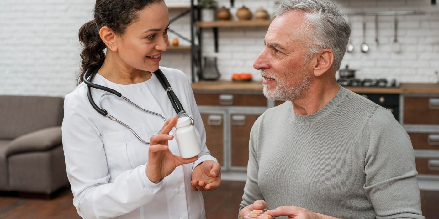 A female doctor shows a pill bottle to an older man in a kitchen, both smiling and talking.