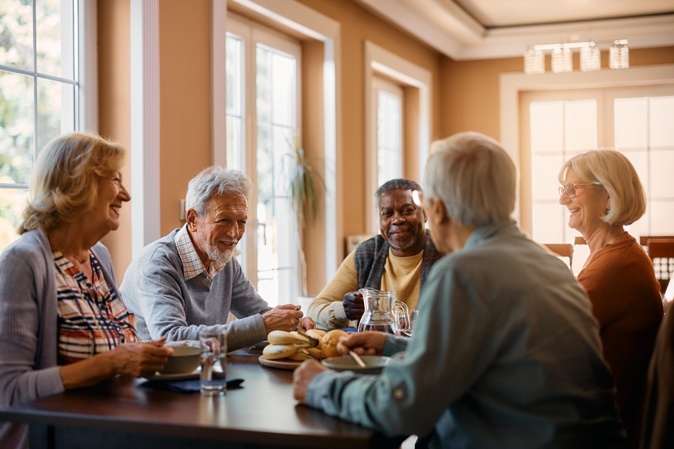 A group of older adults sitting around a table, smiling and talking in a well-lit room.