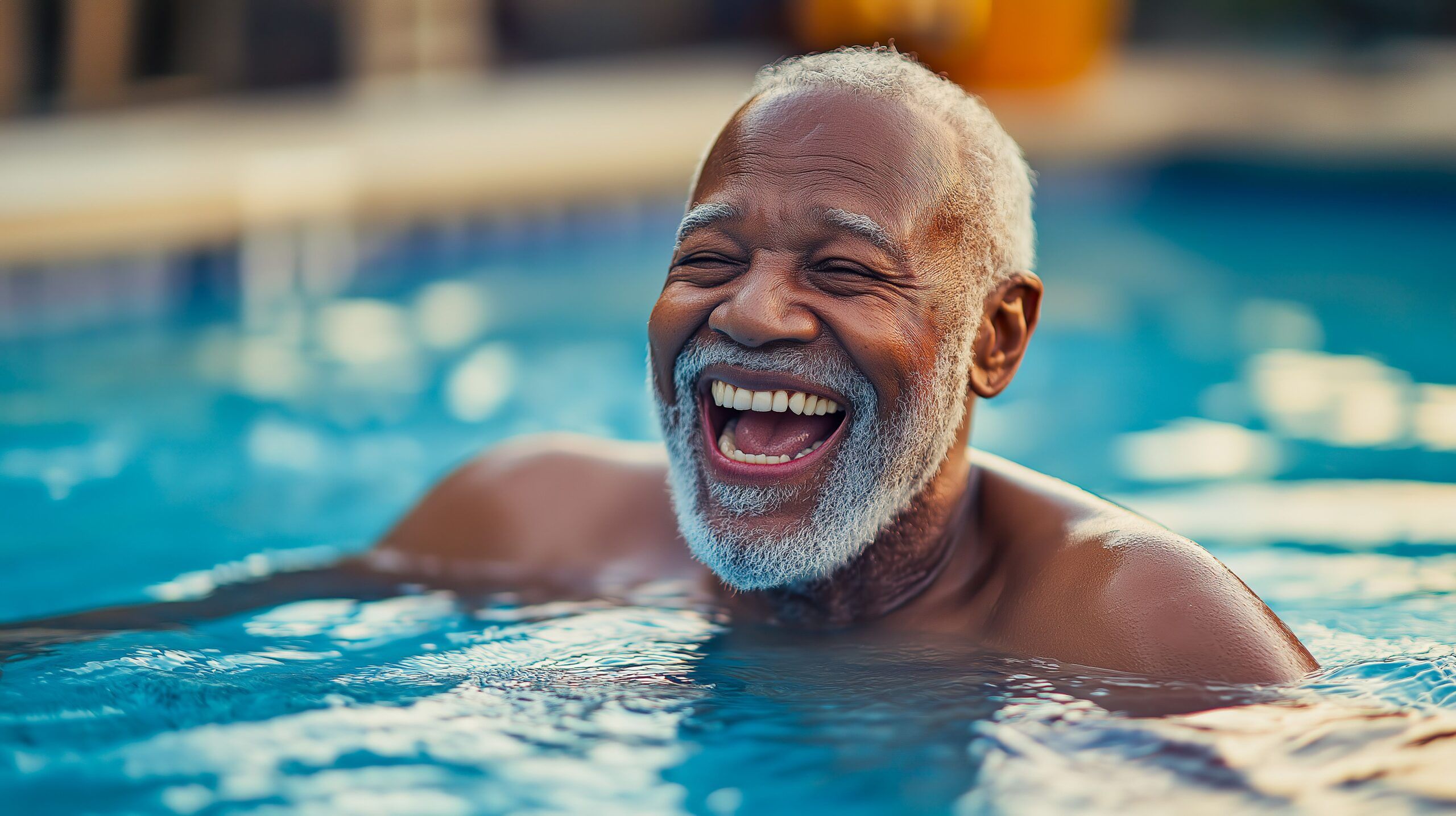 Elderly man with a beard laughing joyfully while swimming in a pool under sunlight.