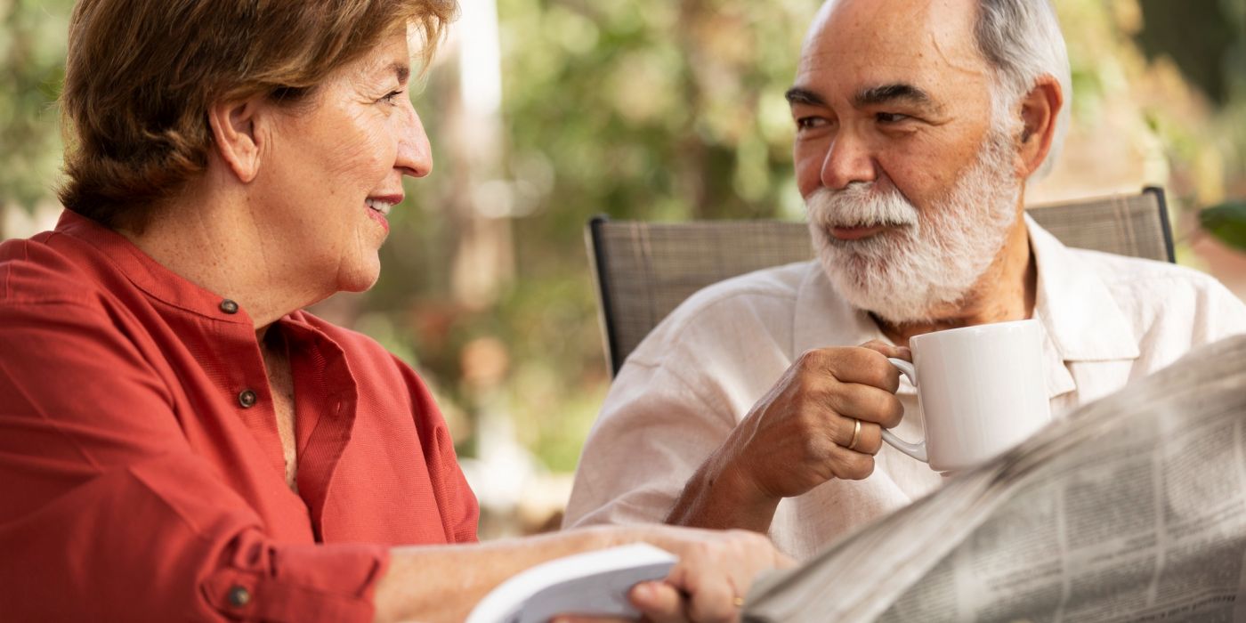 Smiling older couple sits outdoors, drinking coffee and reading a book and newspaper together.