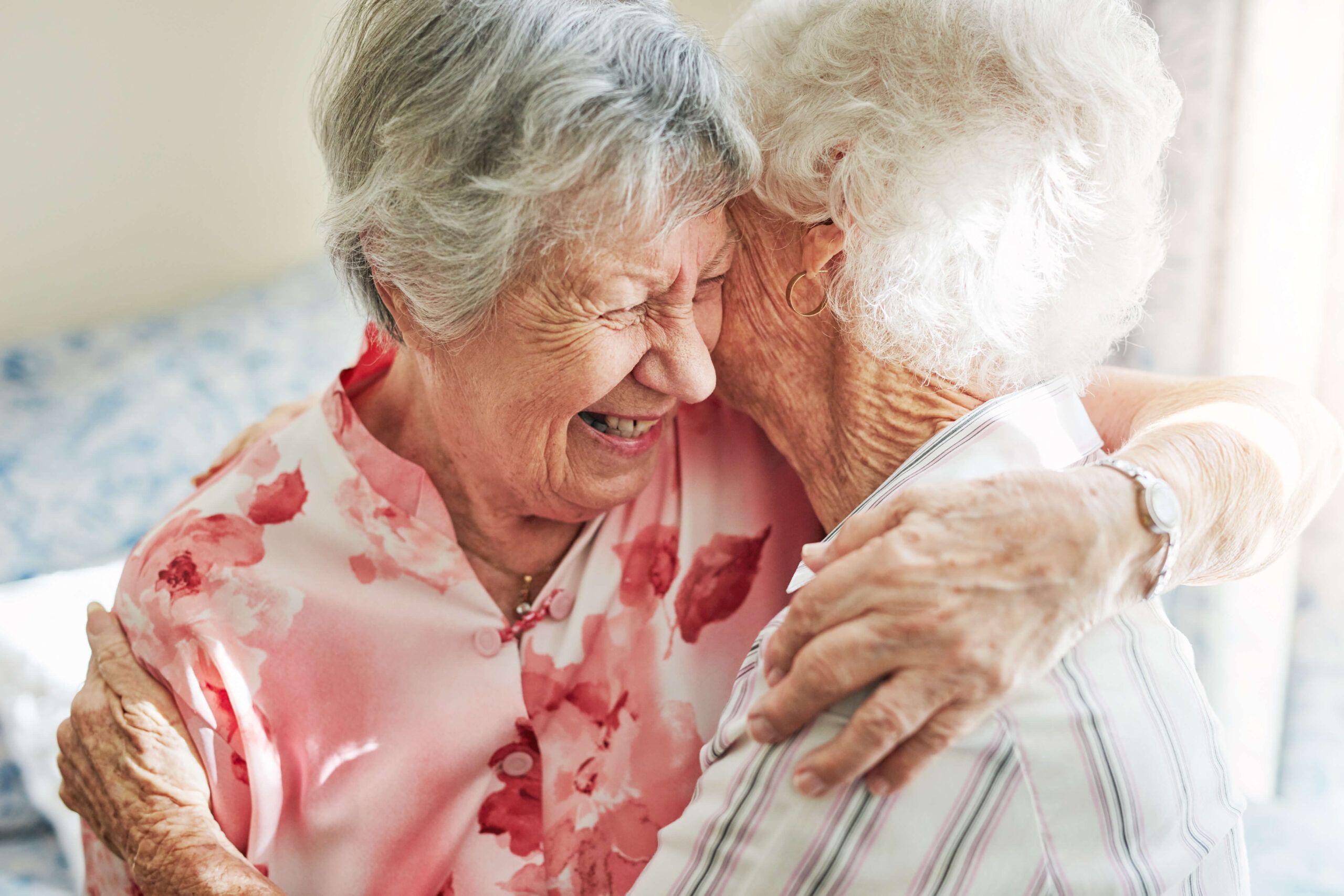 Two elderly women hugging and laughing, wearing floral and striped shirts, in a warmly lit room.