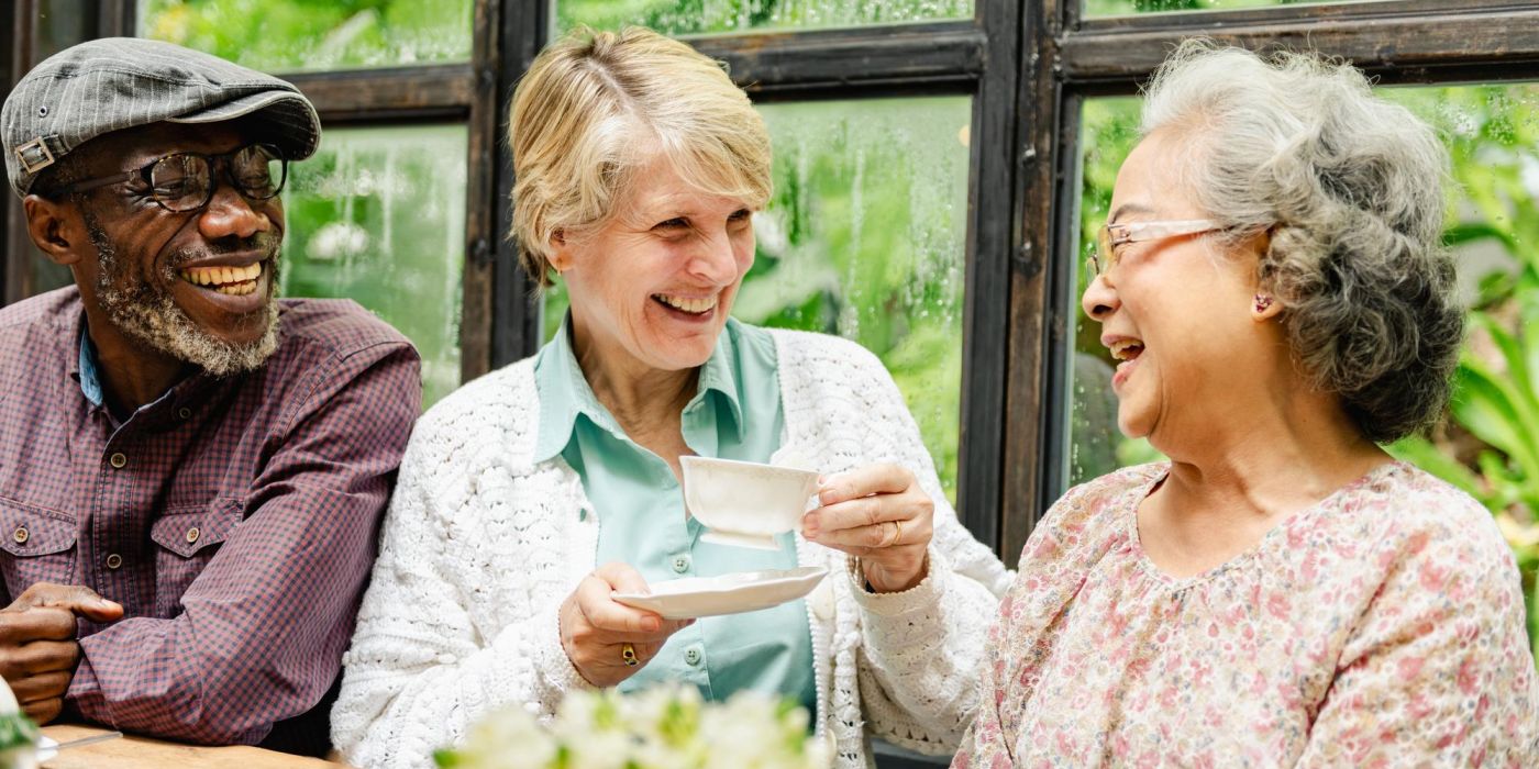 Three older adults laughing and enjoying tea together by a window with greenery outside.