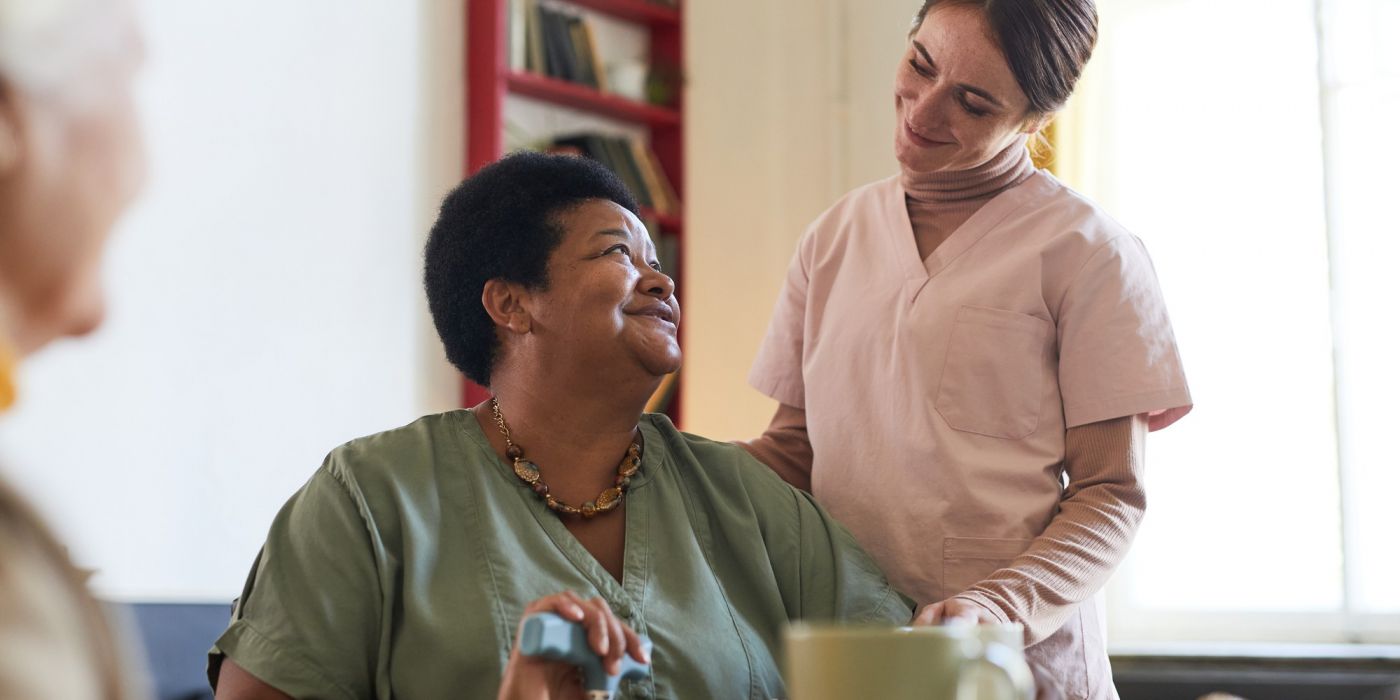 Smiling caregiver stands beside a seated woman, both sharing a warm interaction in a cozy room.
