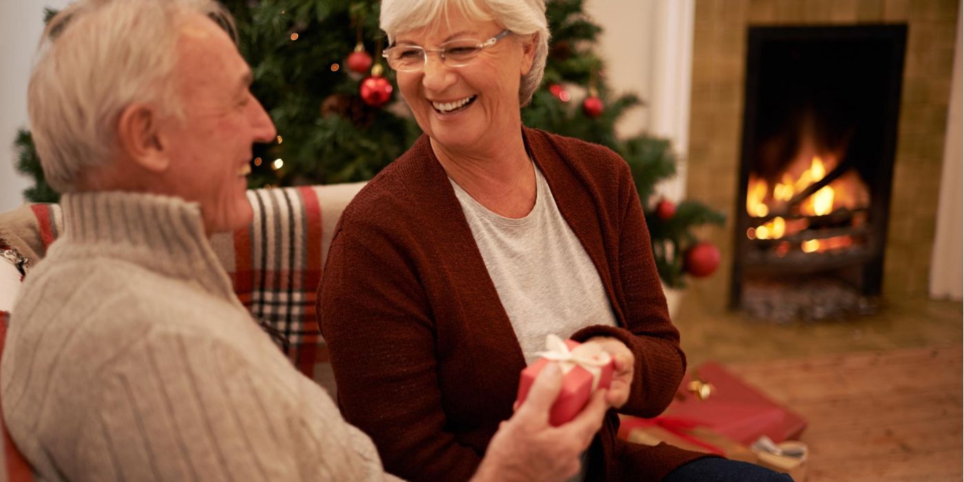 Smiling older couple exchanging a gift by a Christmas tree and fireplace.