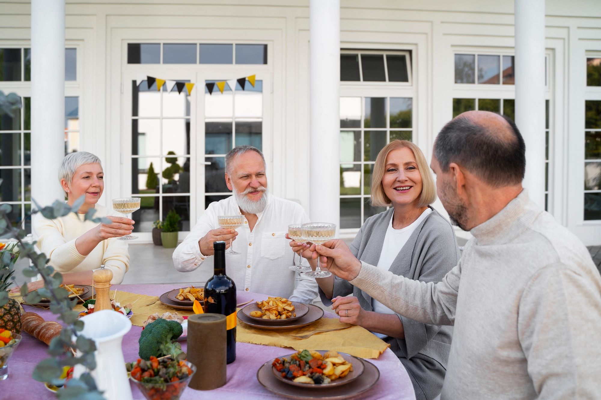 Four older adults sit outdoors at a table, smiling and raising glasses in a cheerful toast.