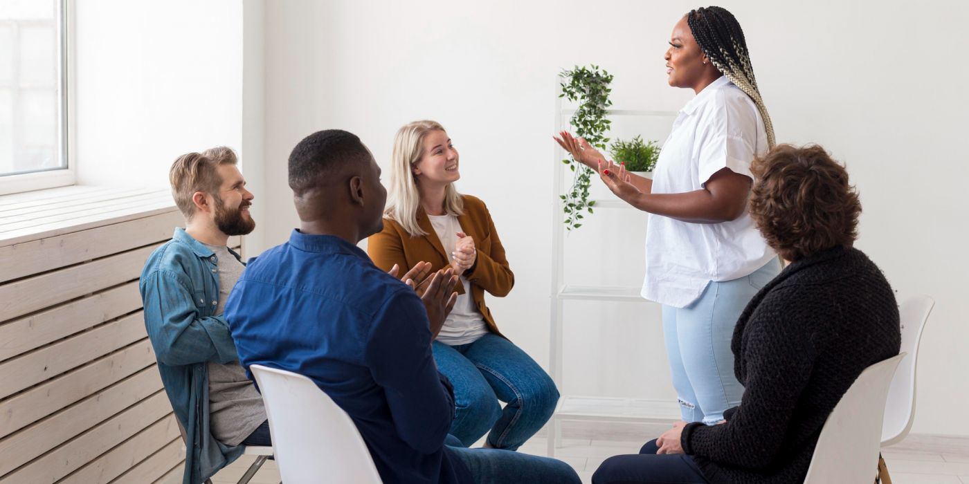 A woman stands speaking to a small group of people sitting in chairs, who are listening and clapping.