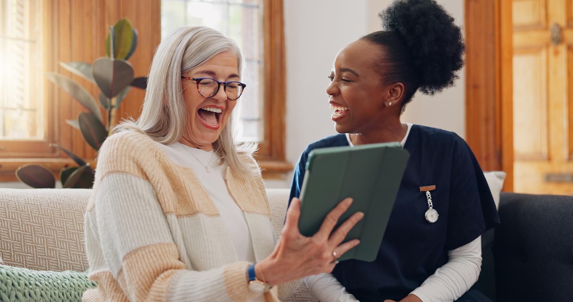 Two women smiling and laughing together while looking at a tablet on a sofa in a bright living room.