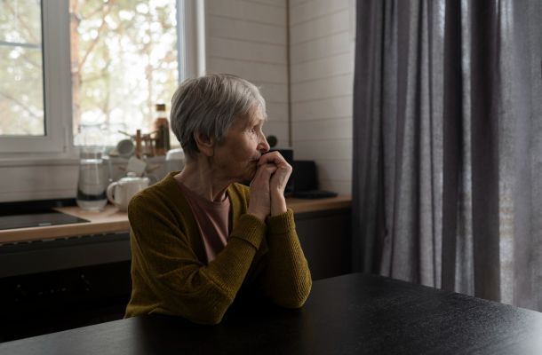 An elderly woman sits at a table, looking thoughtfully out a window in a softly lit kitchen.