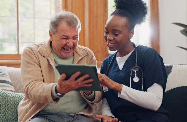 A nurse and an older man smile and look at a tablet together while sitting on a couch.