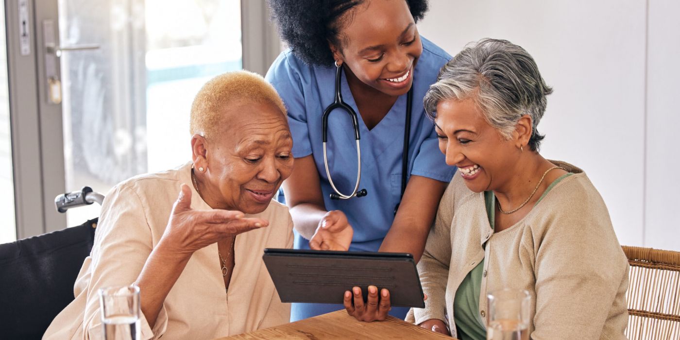 Two senior women smiling at a table with a nurse holding a tablet.