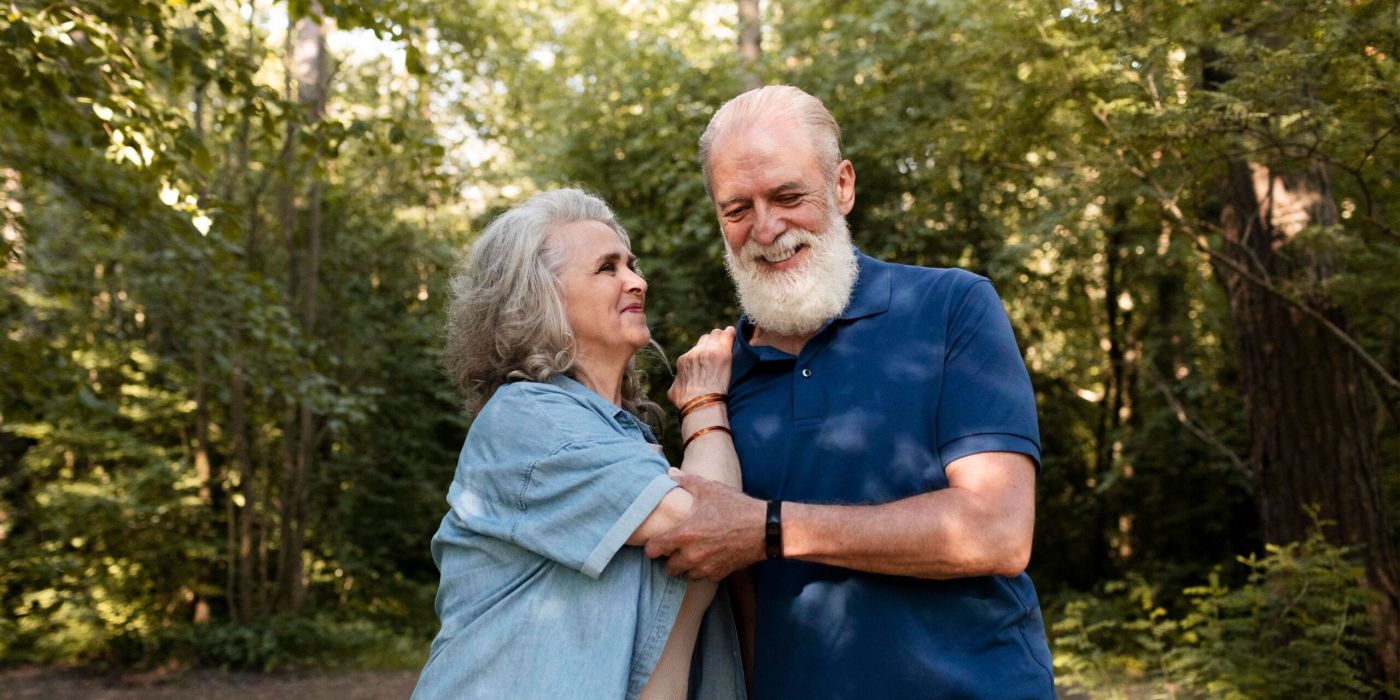 Smiling older couple standing together in a sunlit forest, enjoying each other's company.
