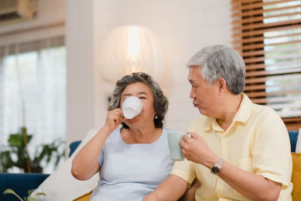 Older couple sitting on a couch at home, enjoying hot drinks and smiling together.