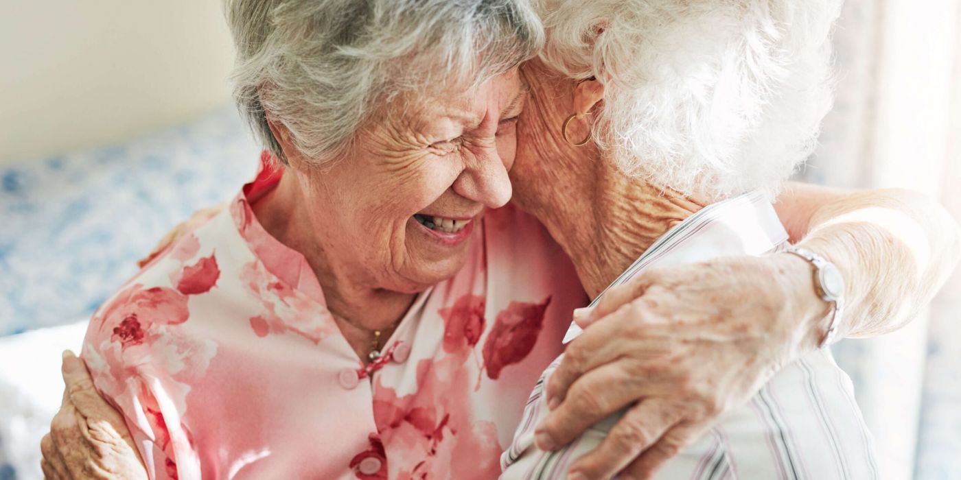Two elderly women hugging and laughing, wearing floral and striped shirts, in a warmly lit room.