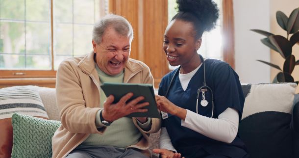 A nurse and an older man smile and look at a tablet together while sitting on a couch.