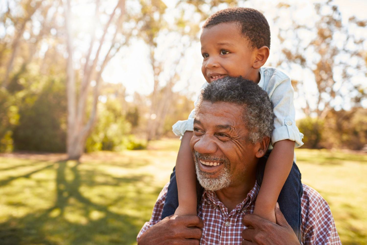 Smiling older man gives a young boy a piggyback ride outdoors on a sunny day in a park.