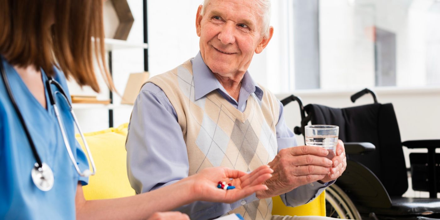 Elderly man holding a glass of water, receiving pills from a nurse, with a wheelchair in the background.