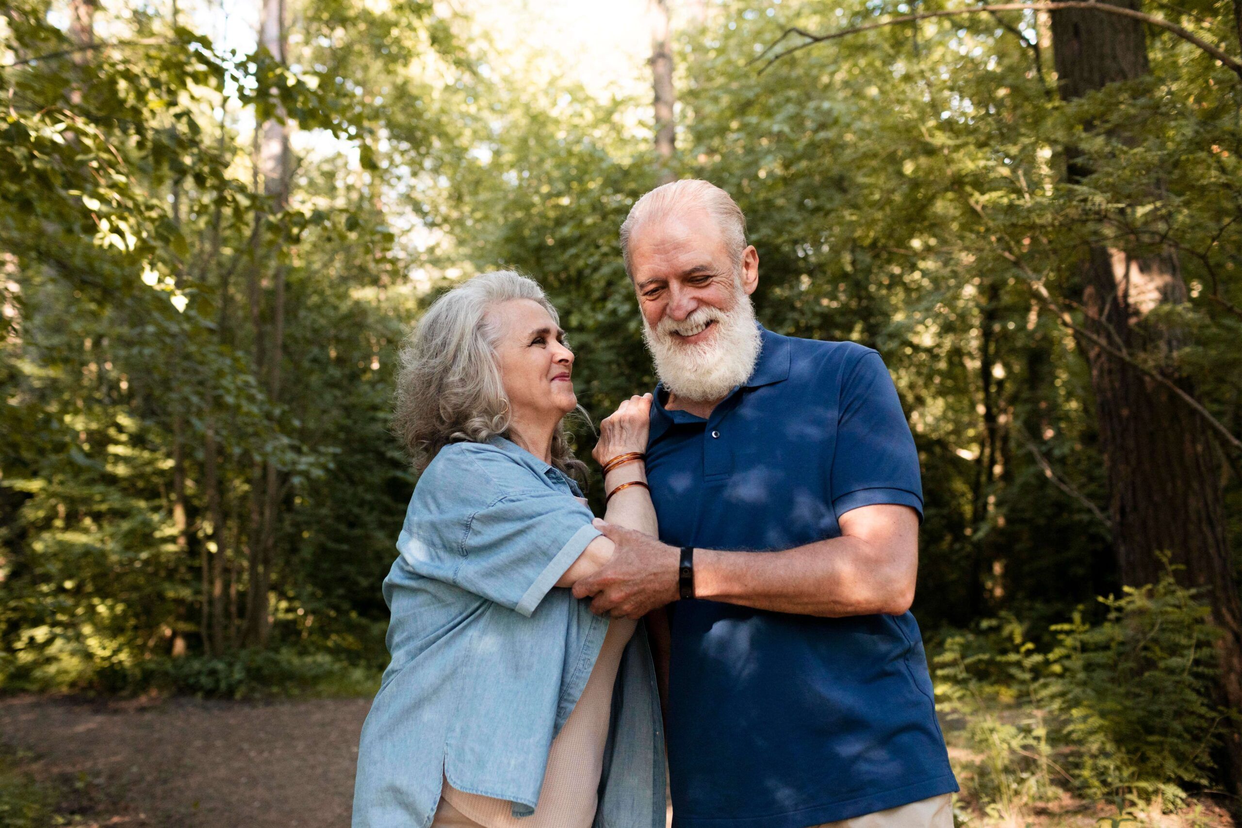 Smiling older couple standing together in a sunlit forest, enjoying each other's company.