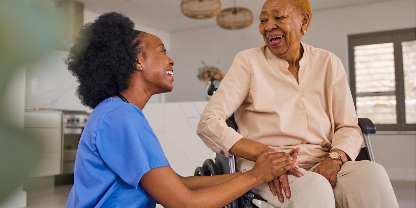 Nurse holding hands and smiling with an older woman in a wheelchair inside a bright home.