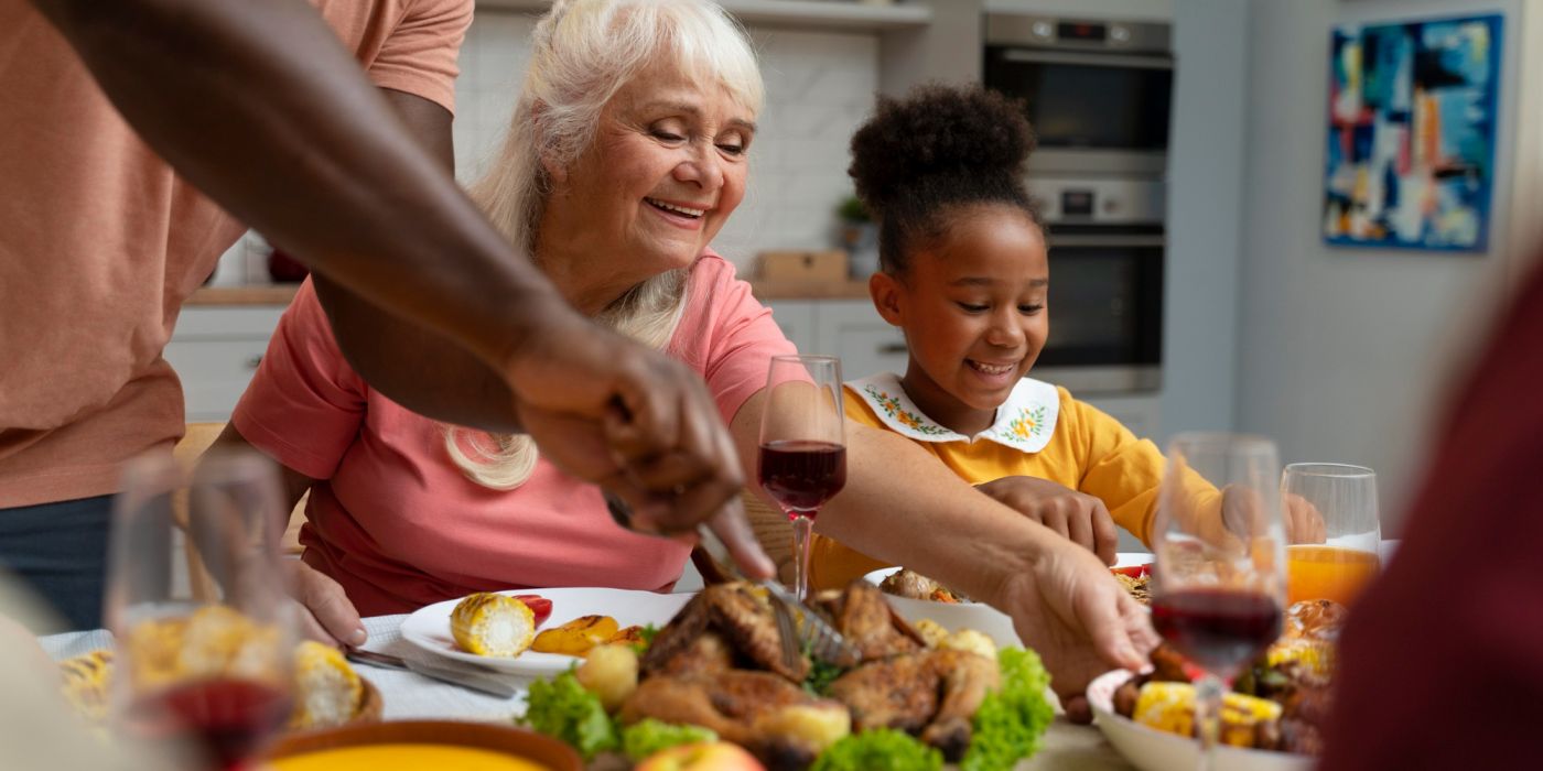 Smiling family sharing a meal at a table, with roast chicken, wine, and various dishes in a cozy kitchen.