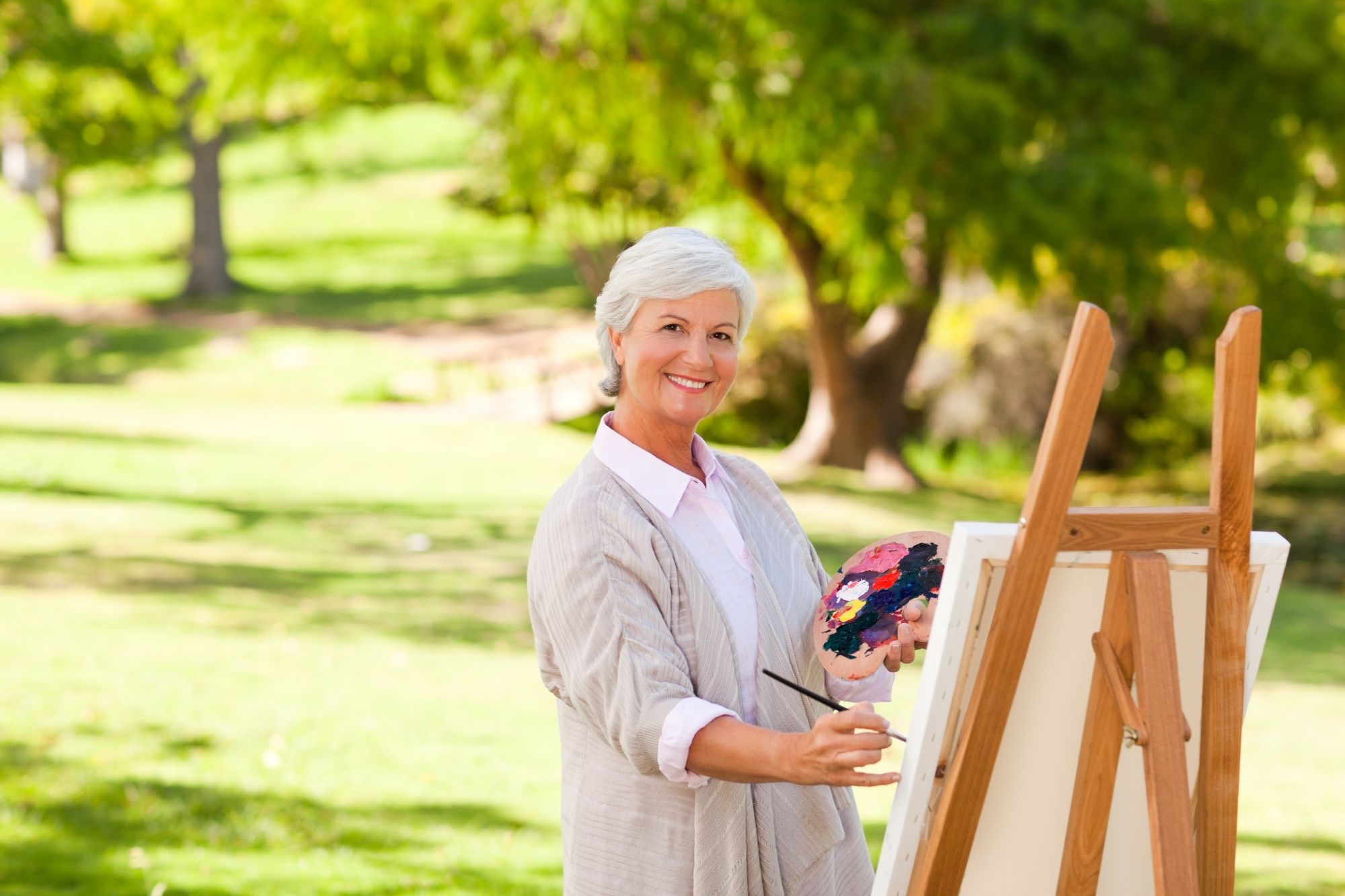Smiling older woman painting at an easel outdoors in a park, holding a palette with colorful paint.