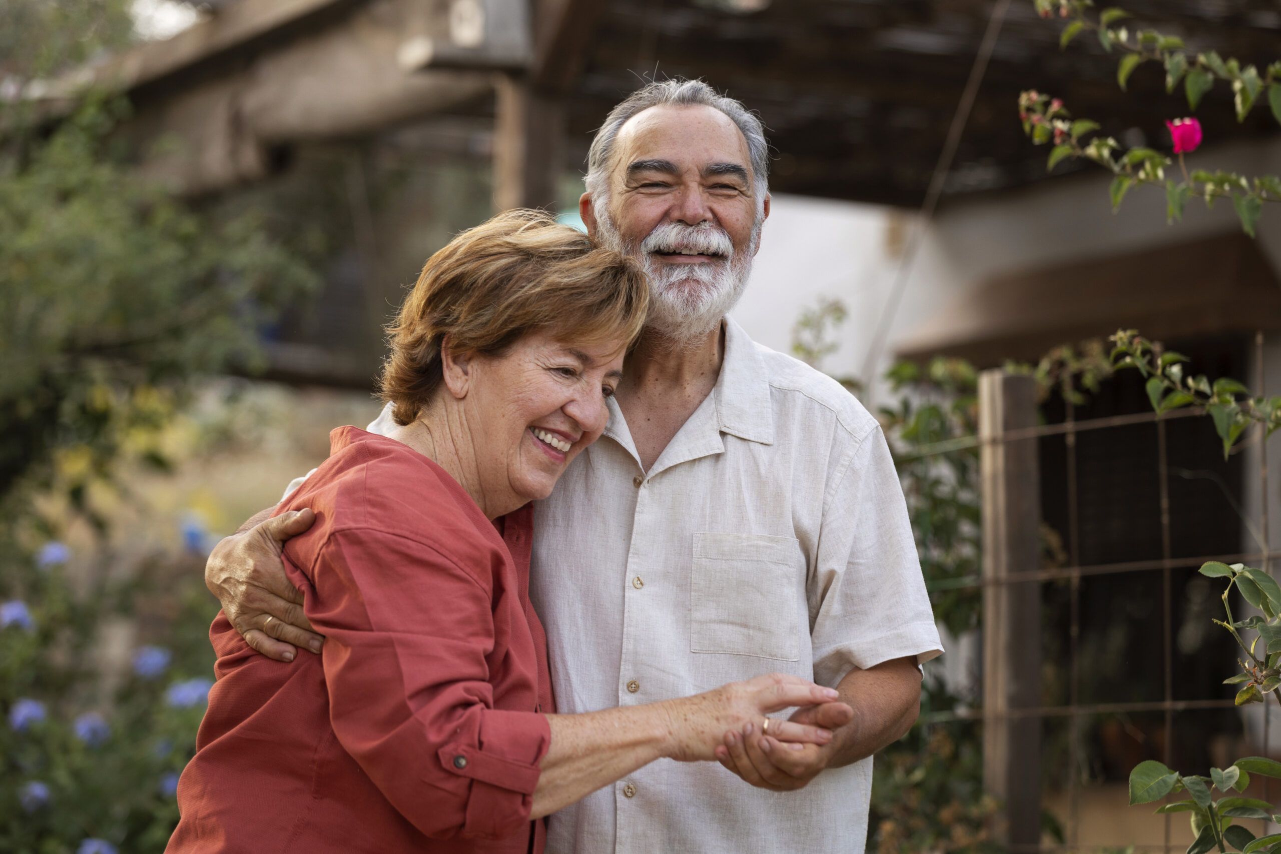 Smiling elderly couple hugging outdoors in a garden, both looking happy and relaxed.