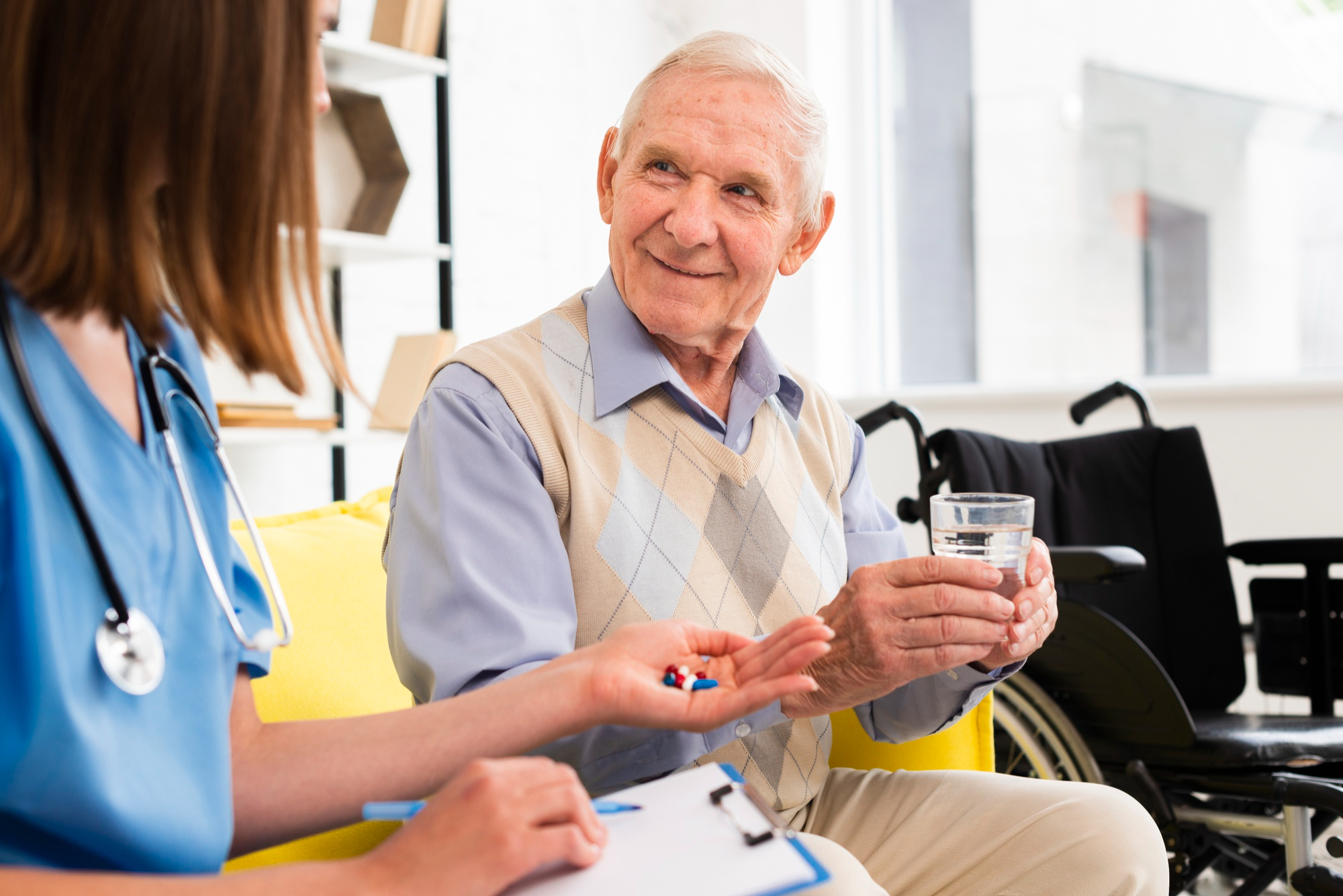 Elderly man holding a glass of water, receiving pills from a nurse, with a wheelchair in the background.
