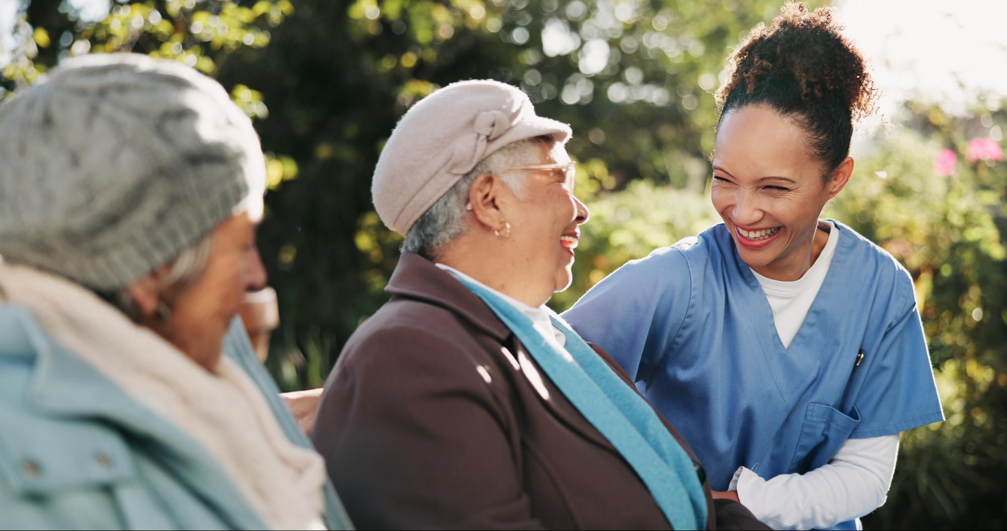 A nurse smiles and talks with two elderly women sitting outside on a bench in a garden.