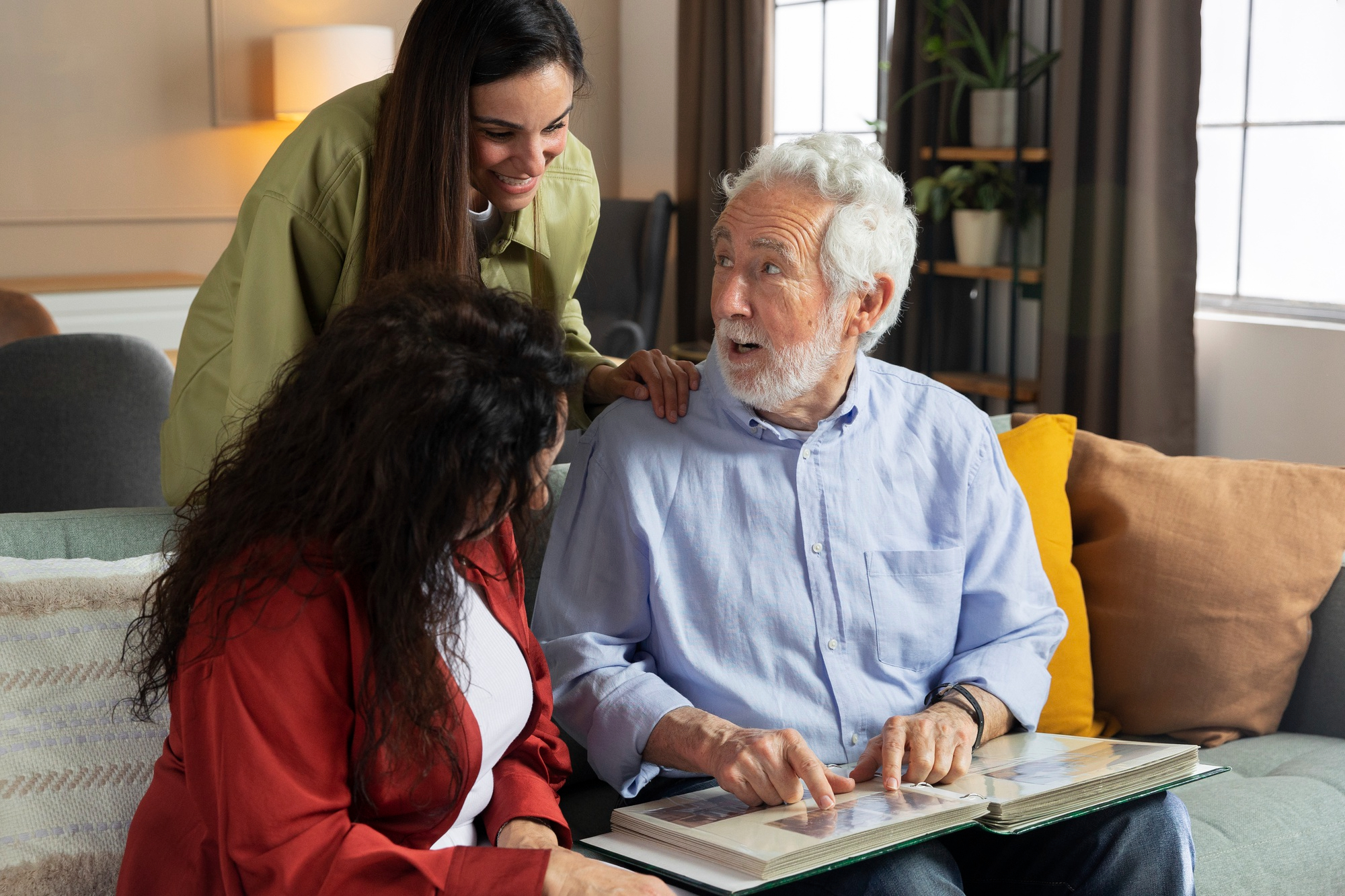 Older man showing a photo album to two women on a couch, all smiling and engaged in conversation.