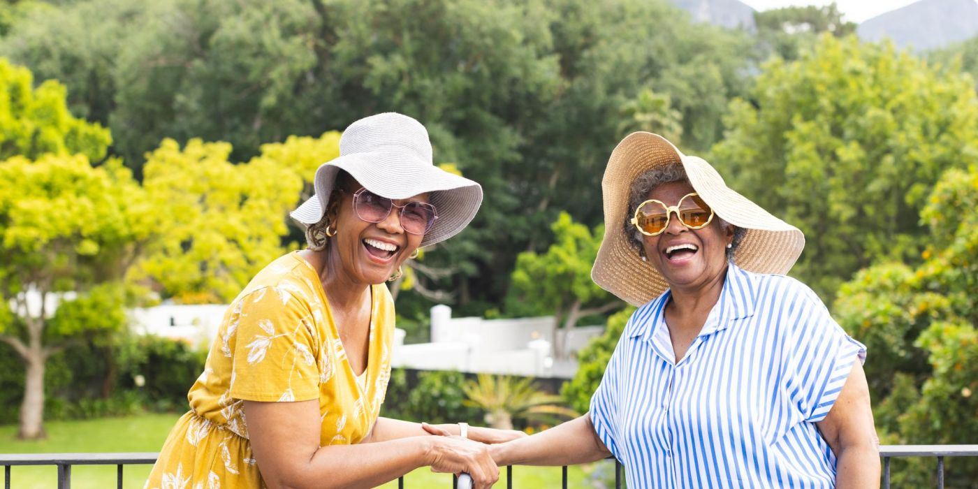 Two older women in sun hats and sunglasses smile and hold hands outdoors with trees in the background.