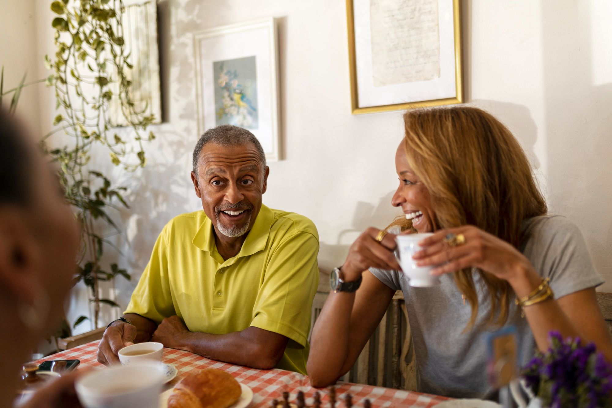 Two people sit at a table, smiling and drinking coffee together in a warmly lit room with framed art on the wall.