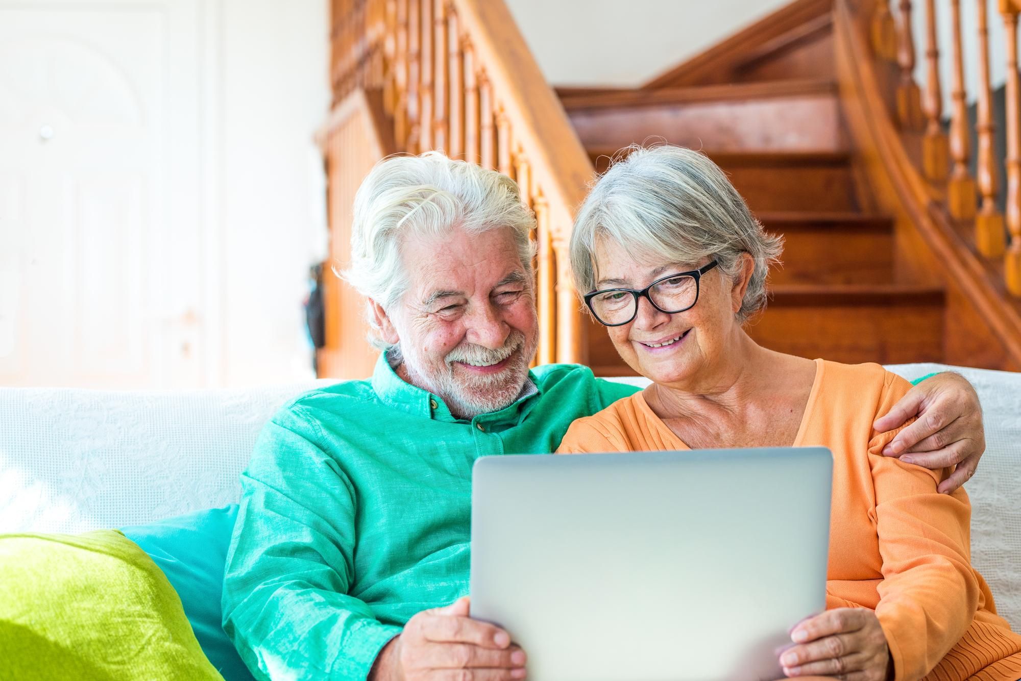 Smiling older couple sitting on a couch, looking at a laptop together in a cozy, sunlit living room with wooden stairs in the background.