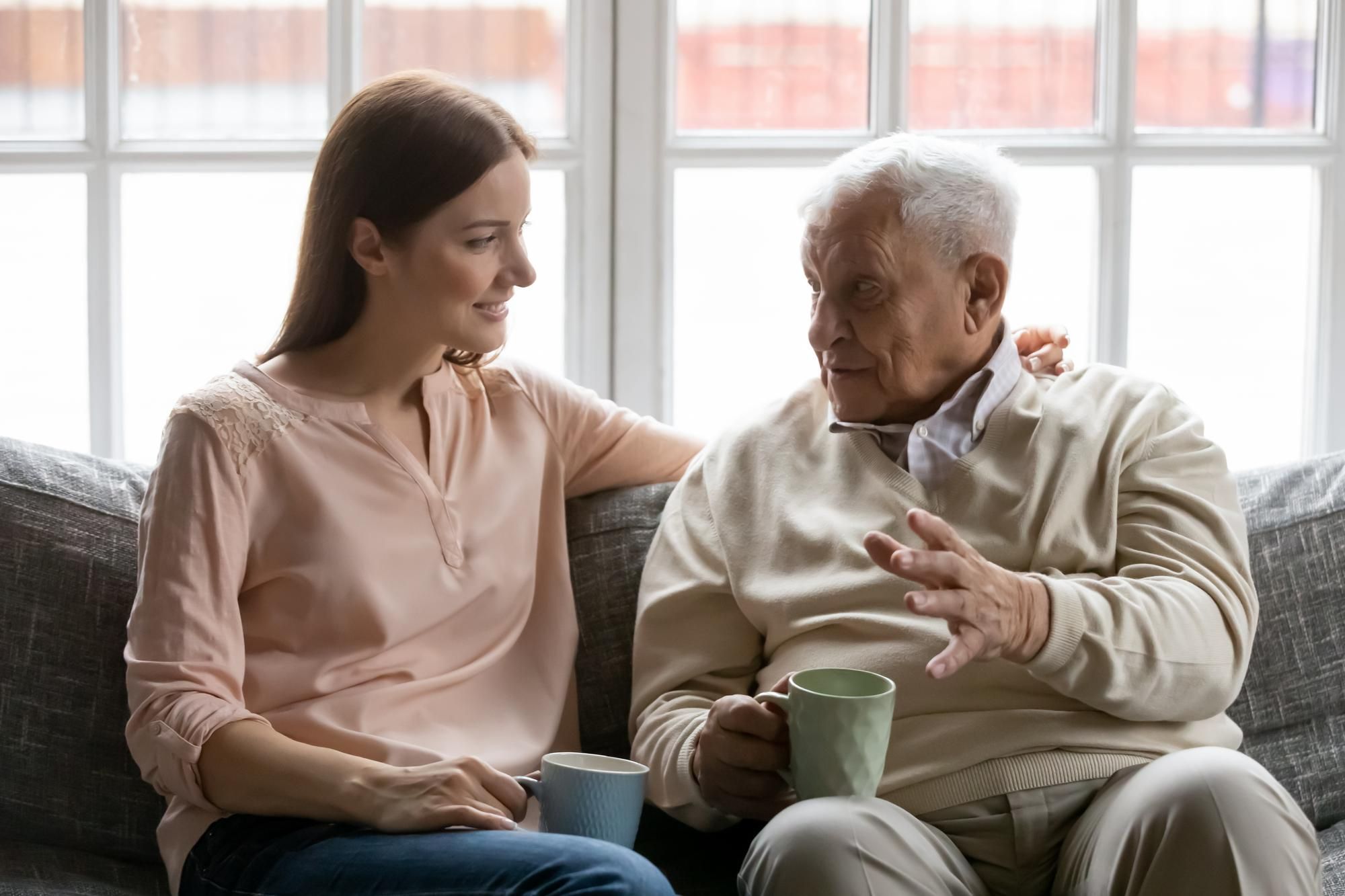 A young woman and an elderly man sitting on a sofa talking while holding mugs.