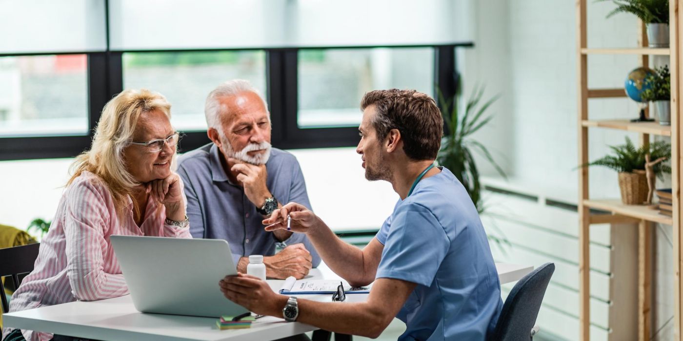 A doctor discusses something with an older couple, using a laptop and documents at a table in an office.