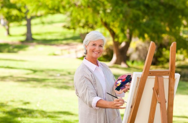 Smiling older woman painting at an easel outdoors in a park, holding a palette with colorful paint.
