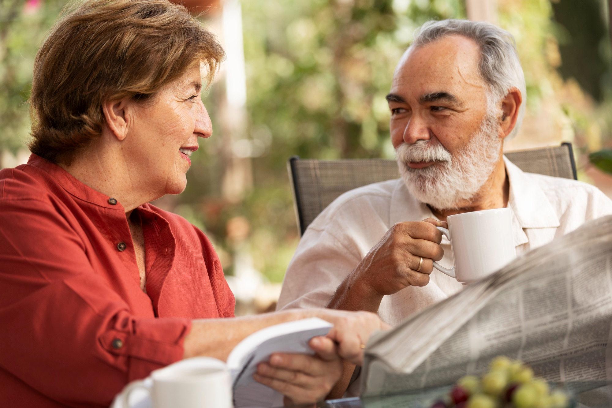 Smiling older couple sits outdoors, drinking coffee and reading a book and newspaper together.