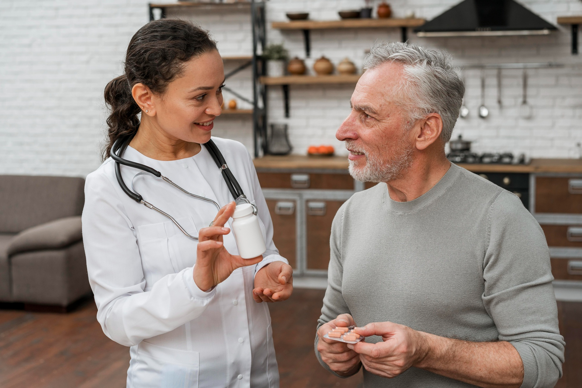 A female doctor shows a pill bottle to an older man in a kitchen, both smiling and talking.