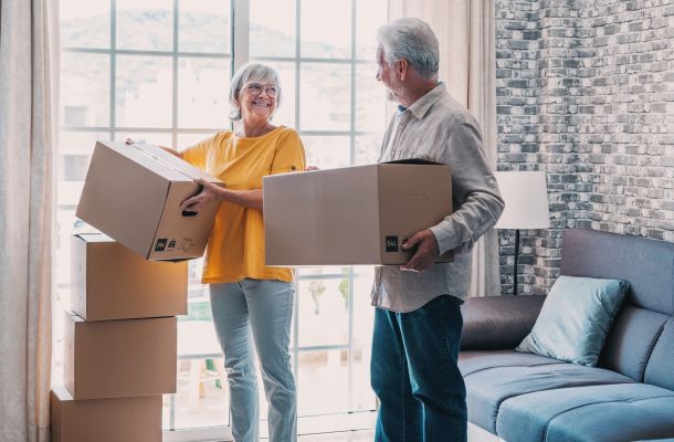 Smiling older couple holding moving boxes in a living room with stacked boxes and a large window.