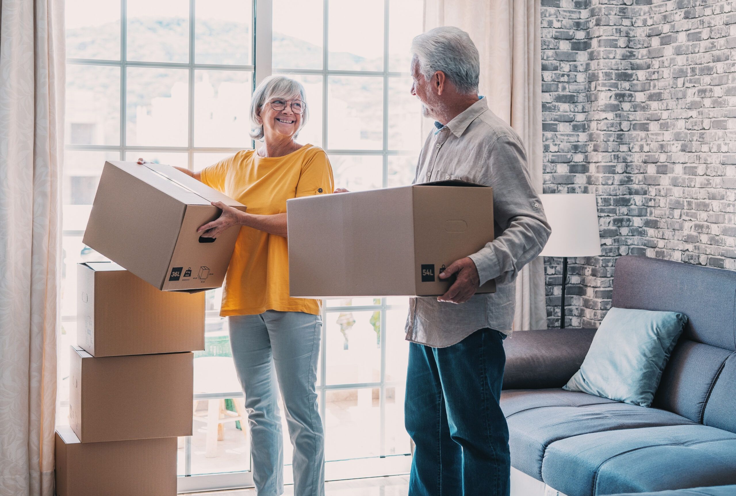 Smiling older couple holding moving boxes in a living room with stacked boxes and a large window.