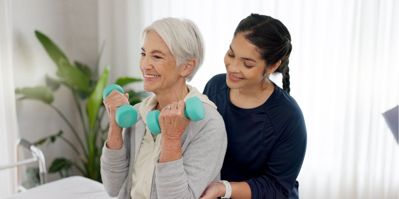 Elderly woman lifts dumbbells with assistance from a caregiver in a bright room.