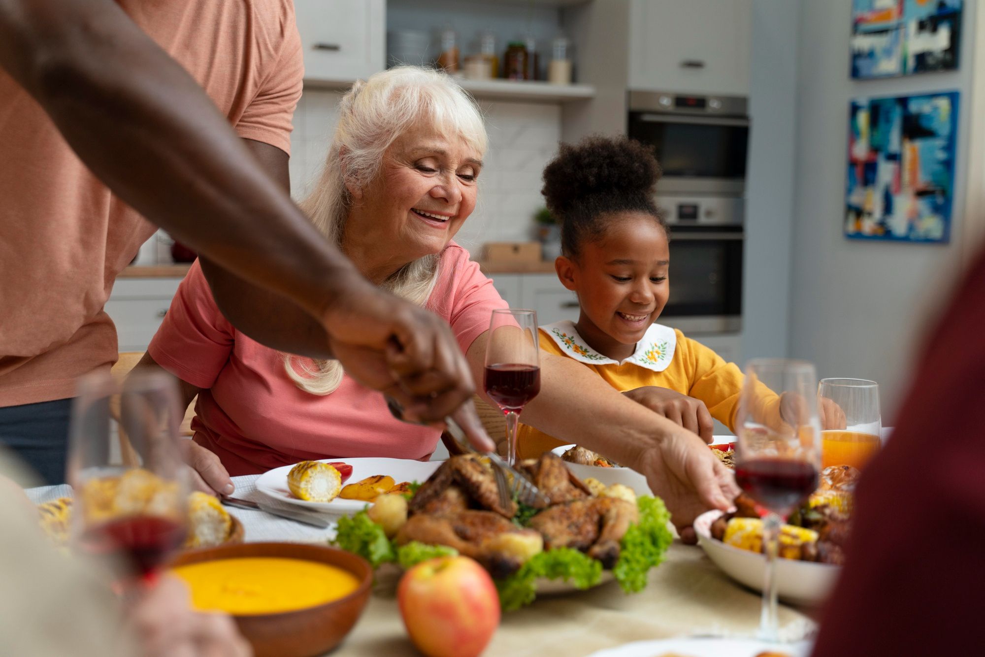 Smiling family sharing a meal at a table, with roast chicken, wine, and various dishes in a cozy kitchen.