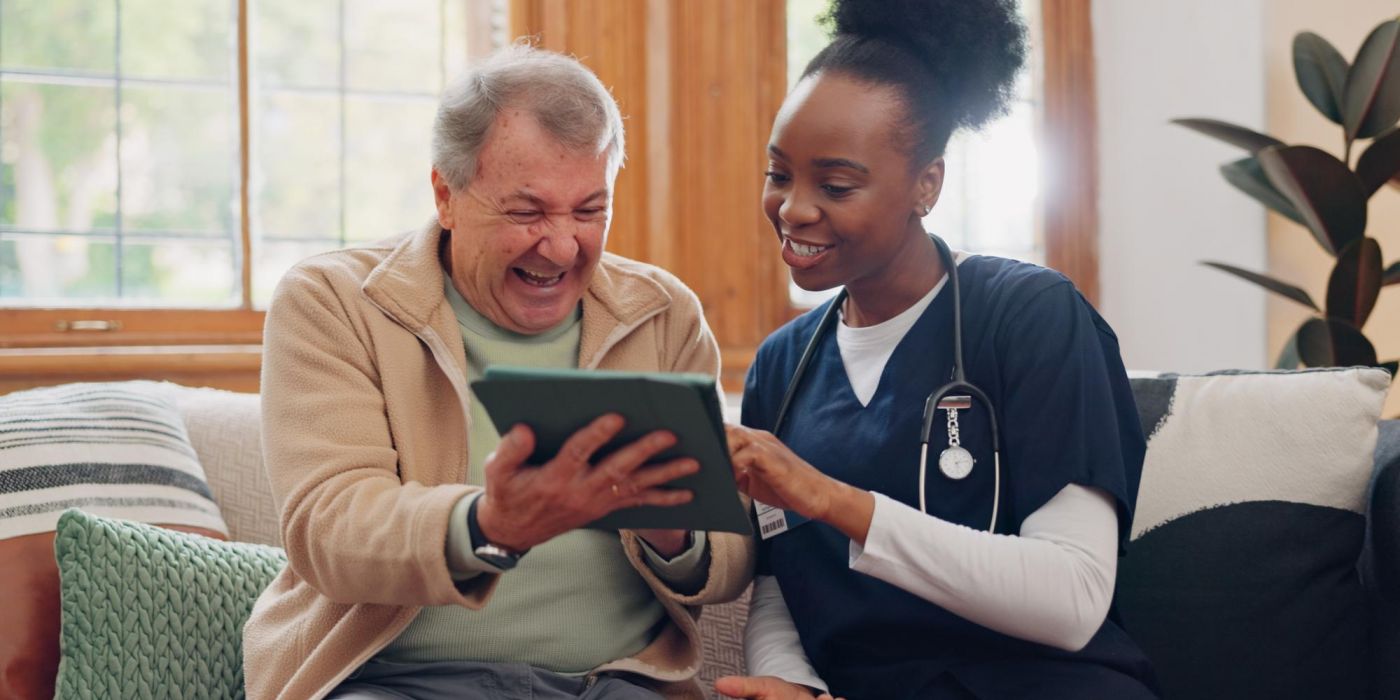 A nurse and an older man smile and look at a tablet together while sitting on a couch.