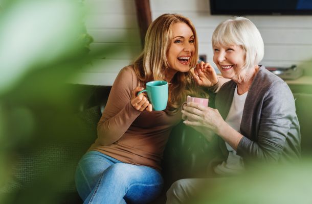 Happy senior mother with adult daughter sitting on couch and holding cups with coffee or tea at home. Togetherness concept