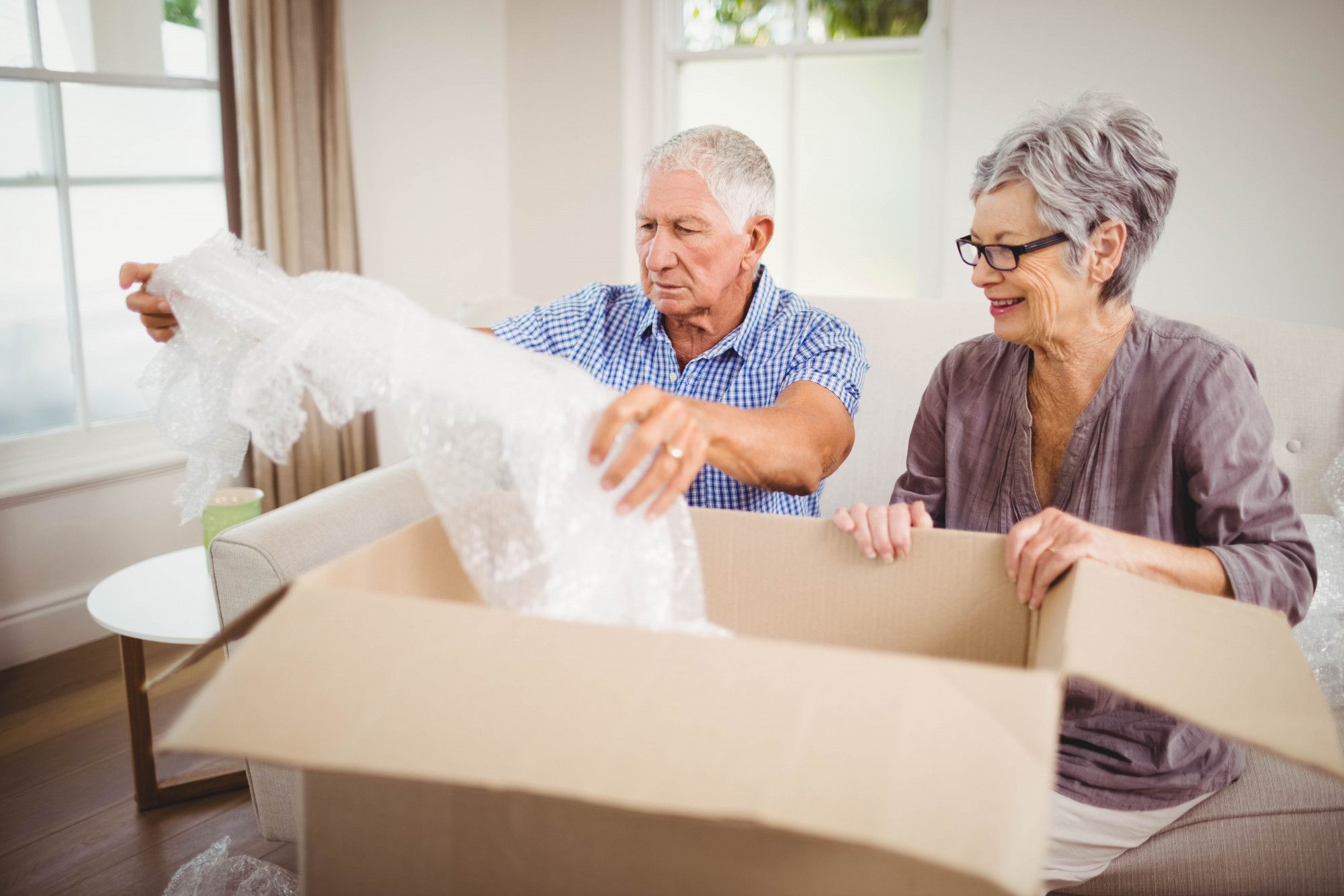 Smiling older couple unpacking a cardboard box and bubble wrap in their living room.