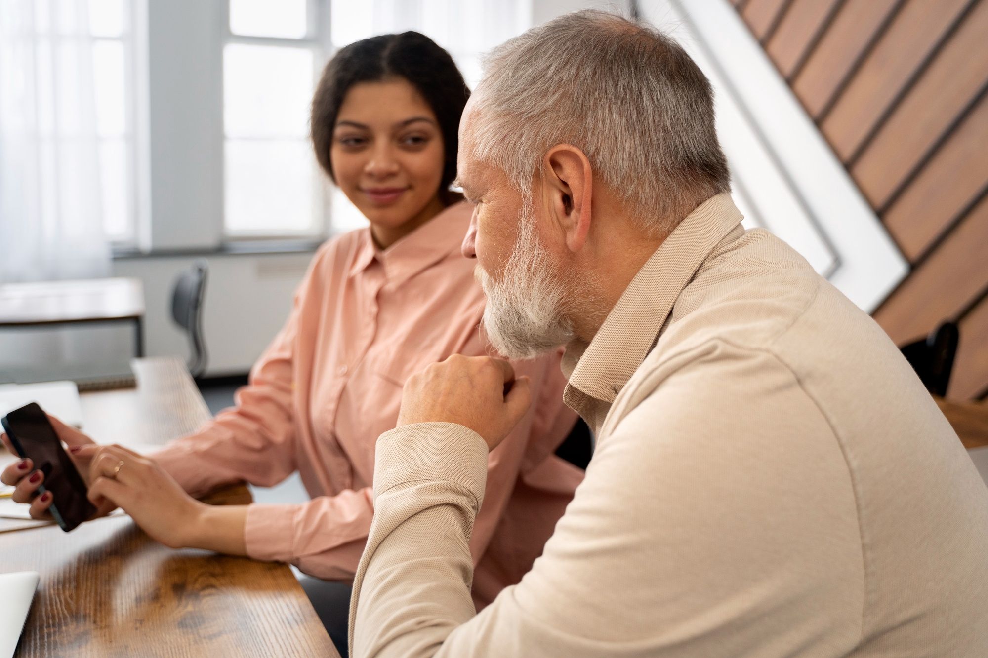 A young woman shows something on her phone to an older man as they sit together at a desk, talking.