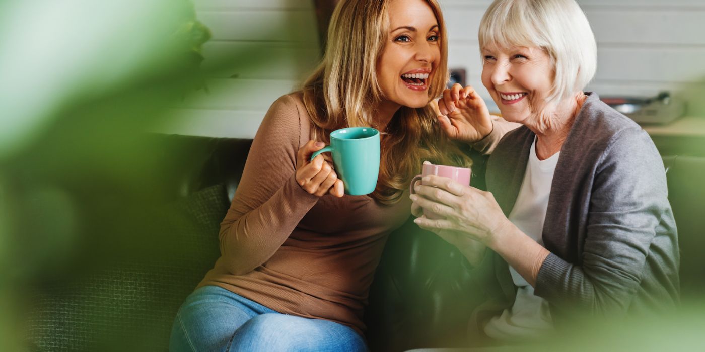 Happy senior mother with adult daughter sitting on couch and holding cups with coffee or tea at home. Togetherness concept