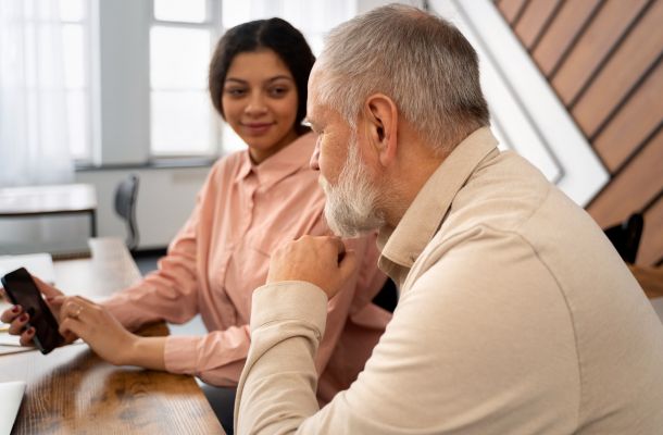 A young woman shows something on her phone to an older man as they sit together at a desk, talking.